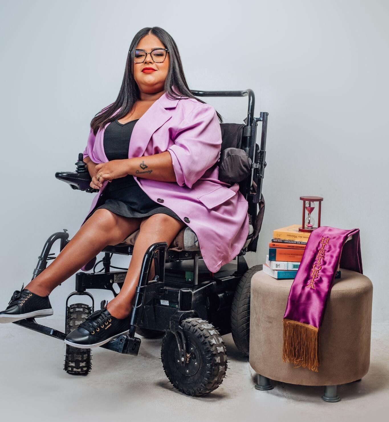 Fotografia em estúdio com fundo branco. No centro da imagem, há uma mulher sentada em uma cadeira de rodas motorizada. Ela tem pele preta, cabelos longos, lisos e escuros, soltos sobre os ombros. Usa óculos de armação preta, batom vermelho e expressão neutra, voltada para a frente. Ela veste um vestido preto curto e um blazer lilás por cima. Calça tênis pretos. As pernas estão descruzadas, e as mãos repousam sobre o colo. À direita da imagem, sobre um puff bege claro, há uma pequena pilha de livros, uma ampulheta e uma faixa de formatura roxa com a palavra "História" bordada em dourado. O ambiente é claro, com iluminação suave e sem outros elementos visuais além dos descritos.